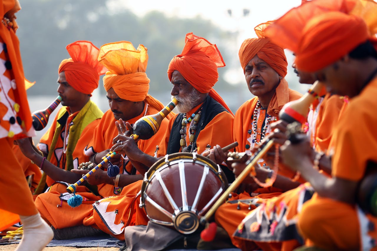 A group of Indian men in vibrant orange traditional attire performing music during a cultural festival outdoors.